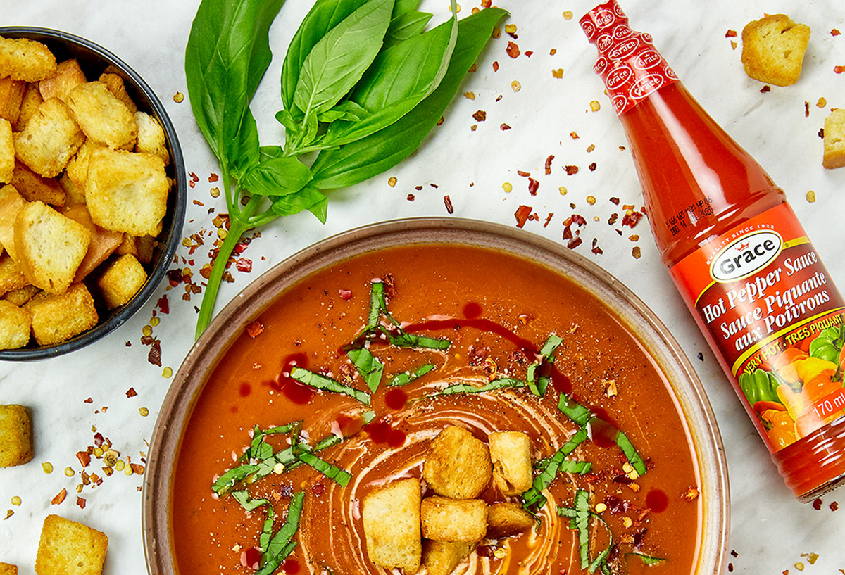 Bowl of soup topped with croutons and basil beside Grace Hot Pepper Sauce and a bowl of croutons