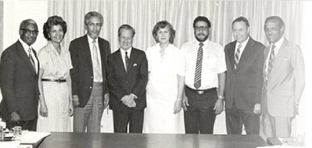 Black-and-white group portrait of eight adults standing together behind a table