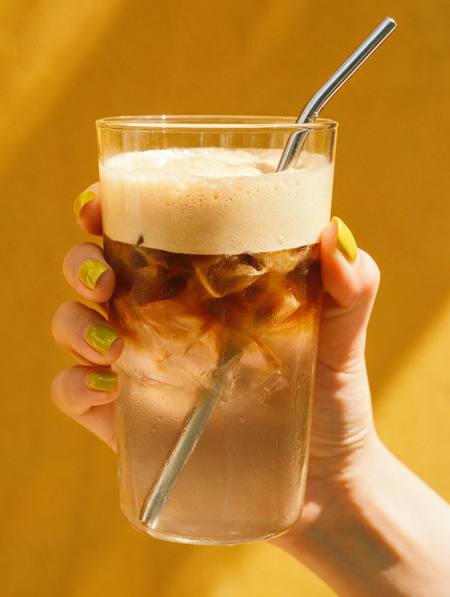 Hand holding a glass of iced coconut water coffee with ice, foam, and a straw