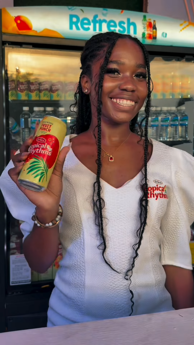 Young woman smiling and holding a can of Tropical Rhythms fruit punch in front of a refrigerated display with a Refresh sign