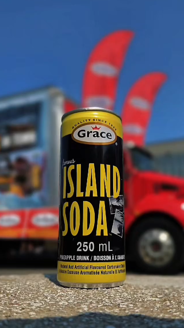 Close-up of 250 mL Grace Island Soda can showing bold ISLAND SODA text, pineapple flavor, blurred red truck and flags