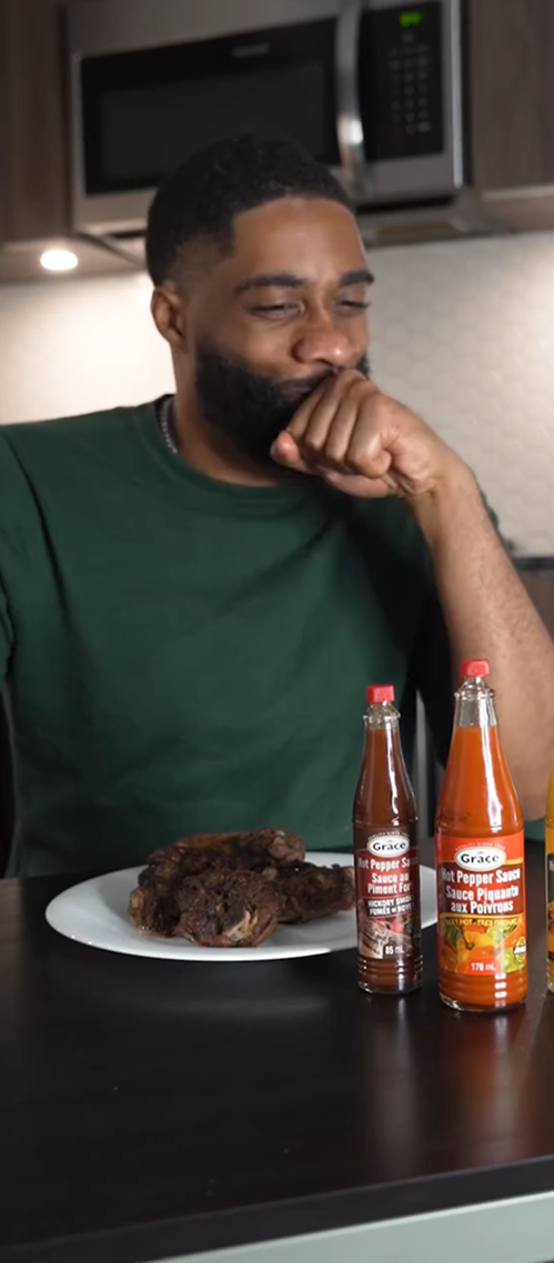 Man laughing at a kitchen island with a plate of jerk chicken and Grace hot pepper sauce bottles in the foreground