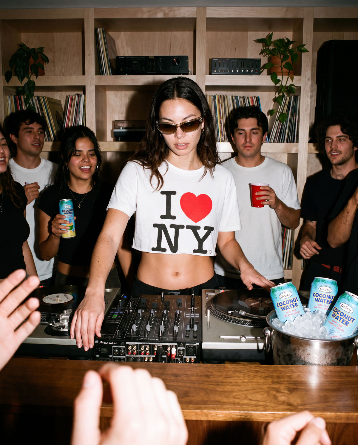 DJ mixing music at a party as friends hold Grace Coconut Water in a record-lined room