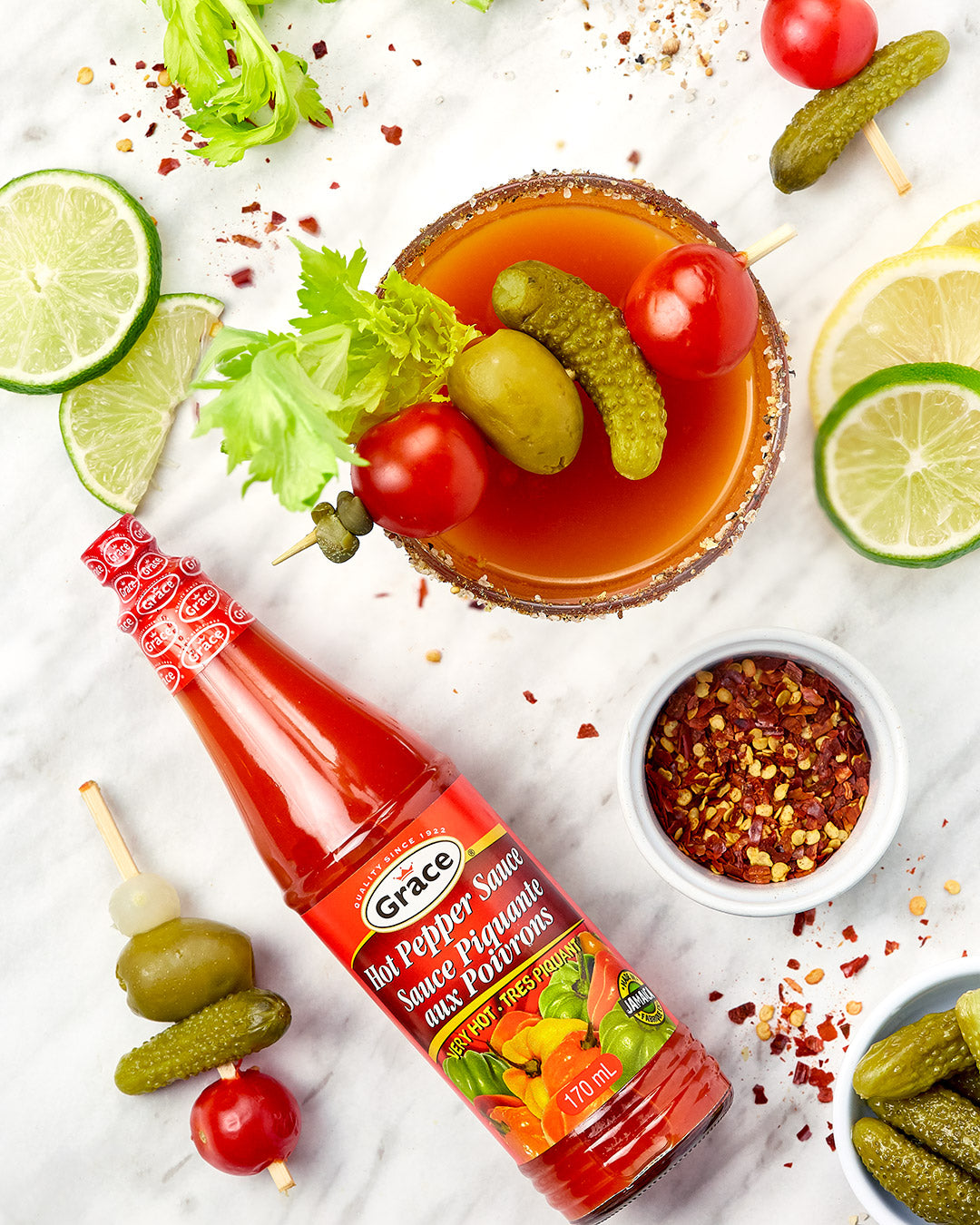 Top-down Grace Hot Pepper Sauce bottle (170 ml) beside a Bloody Mary garnished with pickles, olives and lime slices on marble