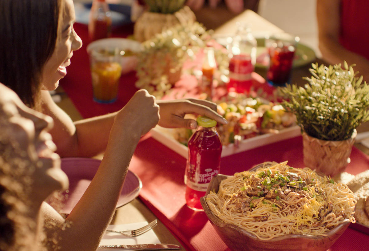 People laughing at a dinner table, one hand reaching for a red bottled drink beside a bowl of noodles on a red tablecloth
