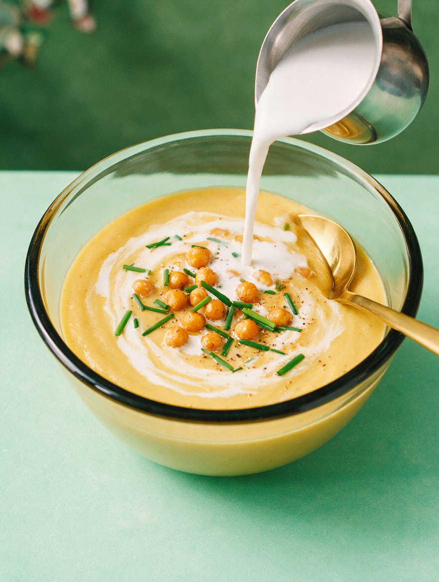 Close up of creamy cauliflower and chickpea soup showcasing chickpeas, chives, and cream being poured on a green background
