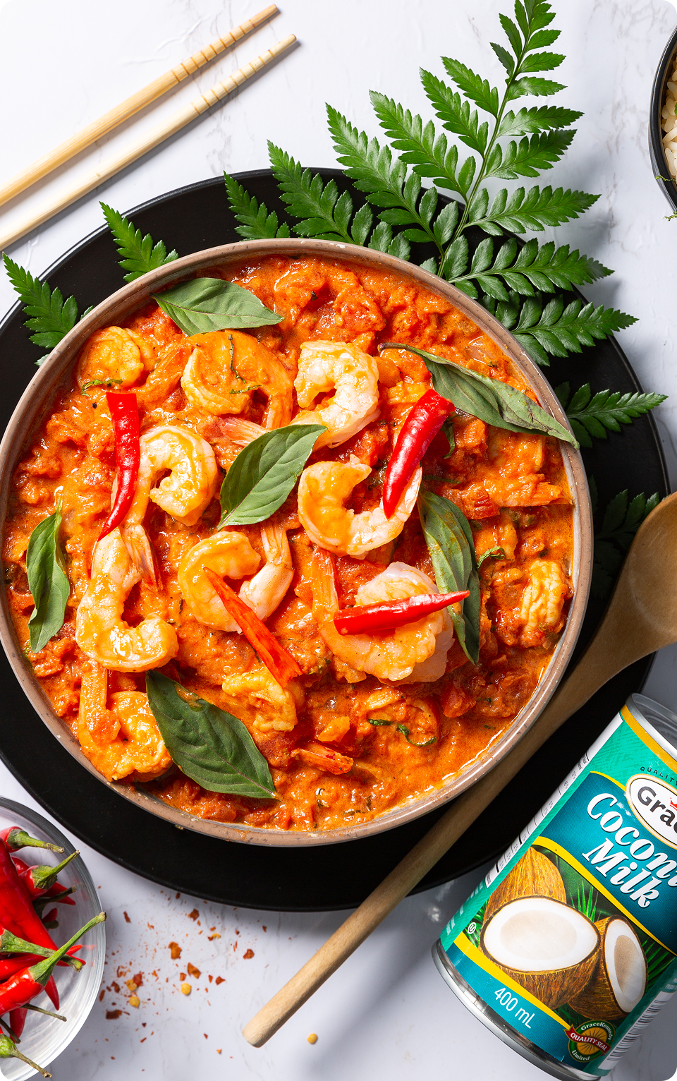 Creamy spicy shrimp curry in a bowl garnished with basil and red chiles, with a can of coconut milk and a wooden spoon